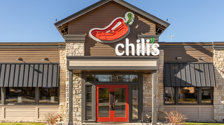 Front exterior of a Chili’s Grill & Bar restaurant featuring the signature red chili pepper logo above the entrance with red double doors and stone-and-wood façade under a clear blue sky.